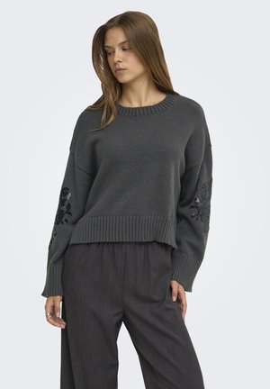 Mujer con un suéter de punto gris oscuro con bordado floral en las mangas y pantalones sueltos gris oscuro, de pie frente a un fondo claro y liso.