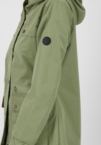 Giacca impermeabile di colore verde chiaro, con cappuccio, bottoni a pressione, tasche laterali e un patch con logo nero sulla parte superiore del braccio. Texture liscia.