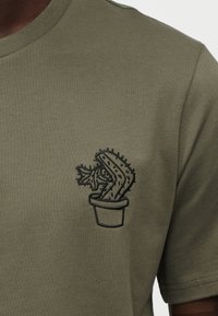 Λαδί πράσινο T-shirt με σχέδιο κεντητού μαύρου κάκτου σε γλάστρα στην αριστερή πλευρά του στήθους. Κοντά μανίκια, στρογγυλή λαιμόκοψη, μαλακή υφή.