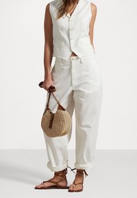 Kvinna i vit ärmlös knäppt väst, högmidjade vita uppvikta byxor, håller i en vävd handväska och har bruna sandaler med remmar.