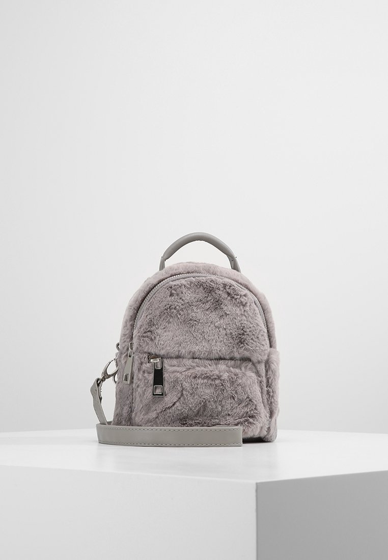 Petit sac à dos gris en fausse fourrure avec poche avant zippée, poignée supérieure et sangle d'épaule réglable, présenté sur une surface blanche.