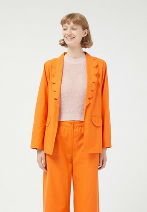 Blazer de lino naranja con solapas onduladas y dos bolsillos frontales, combinado con una camiseta de punto texturizada en rosa claro y pantalones naranja a juego.