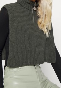 Top senza maniche di colore verde oliva in maglia con collo alto, dettaglio della cerniera frontale e texture a coste, abbinato a pantaloni aderenti di colore verde chiaro.