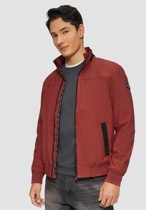 Veste légère rouge avec un col haut, fermeture éclair avant et détails en accent noir. Le matériau semble lisse avec une finition légèrement brillante.