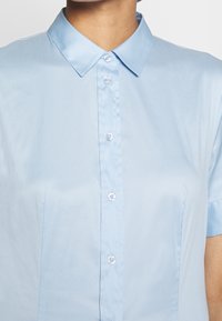 Personne portant une chemise bleu clair à manches courtes boutonnée avec un col pointu, montrée du cou au haut du torse.