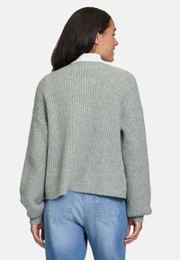 Femme de dos portant un pull en tricot épais gris clair avec des épaules tombantes et un jean bleu clair, devant un fond uni.