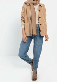 Beige manchesterjacka med två framfickor, bärs över en ljus tröja, kombinerad med blå jeans och bruna ankelstövlar. En brun scarf är draperad runt halsen.