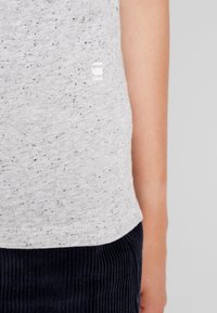 Närbild av en person som bär en ljusgrå fläckig t-shirt med en liten vit "RAW"-logotyp och mörka ribbade byxor.