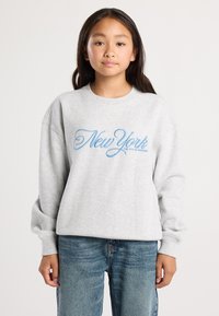 Nuori tyttö, jolla on päällään vaalea harmaa huppari, jossa lukee "New York" sinisellä käsin kirjoitetulla fontilla, sekä väljät siniset farkut. Hän seisoo tasaisella taustalla.