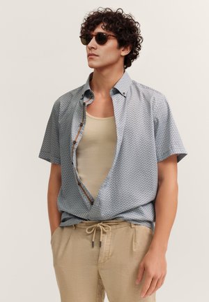 Junger Mann mit lockigem Haar und Sonnenbrille, der ein gemustertes Kurzarmhemd über einem beigen Tanktop und beigen Hosen mit Kordelzug trägt.