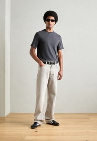T-shirt gris à manches courtes, jean large beige, ceinture noire, baskets noires avec rayures blanches et lunettes de soleil. Textures lisses, style minimaliste.