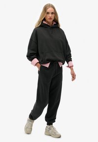 Sort oversized hoodie med en frontlomme, lagd over en lyserød skjorte. Kombineret med matchende sorte joggerbukser og beige sneakers.
