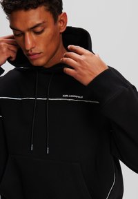 Sweatshirt à capuche noir avec des coutures blanches. Dispose d'une poche avant, de cordons de serrage réglables, et du logo "KARL LAGERFELD". Matériau en mélange de coton.