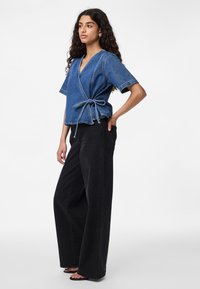 Top avvolgente in denim blu con maniche corte e dettaglio a fiocco in vita, abbinato a pantaloni neri a gamba larga e sandali flat.