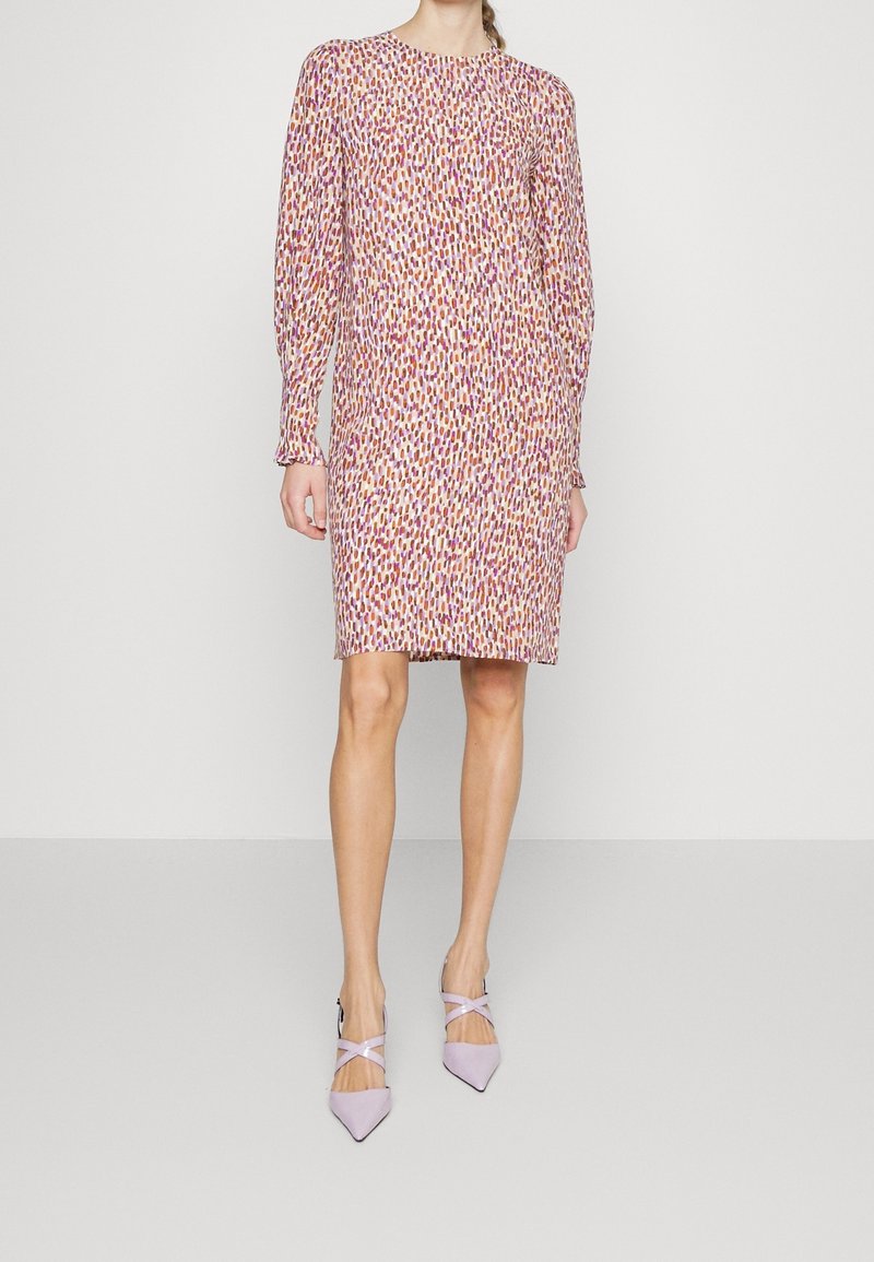 Femme portant une robe longue manche longue jusqu'aux genoux avec des taches abstraites roses, orange et violettes et des escarpins lavande à bout pointu avec des brides à la cheville.