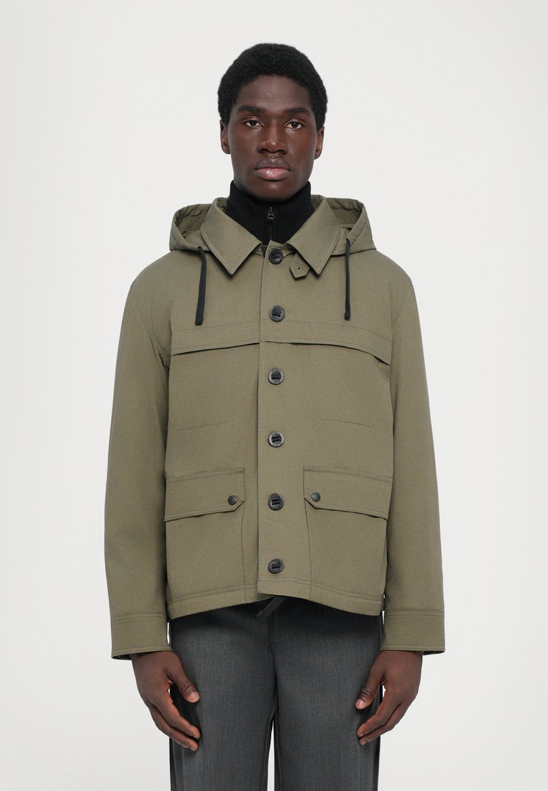 Olivgrüne Kapuzenjacke mit Knopfleiste, zwei Brusttaschen und verstellbaren Kordeln. Der Stoff wirkt glatt und leicht strukturiert.