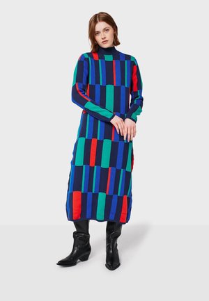 Frau, die ein langes, mehrfarbiges, geometrisch gemustertes Kleid mit vertikalen Streifen in Blau, Grün, Rot und Schwarz trägt, kombiniert mit schwarzen Stiefeln.