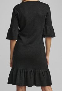 Svart klänning med figurnära liv, utsvängda trekvartsärmar och volangkant. Tillverkad av ett texturerat material med subtil lyster.