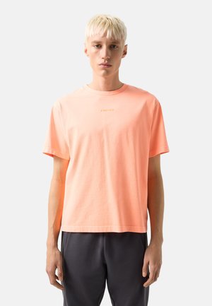 Junger Mann mit kurzen blonden Haaren trägt ein helloranges "FIRE+ICE" T-Shirt und dunkle Hosen, steht vor einem schlichten weißen Hintergrund.