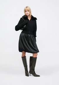 Pulover negru ajustat cu fermoar și guler din blană artificială, asortat cu o fustă balon din satin negru și cizme maro din piele cu toc până la genunchi.