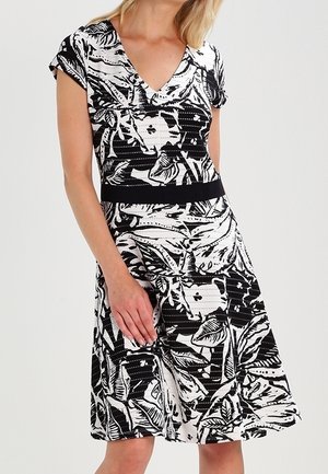 Kvinde iført en knælængde kjole med sort og hvidt blomstermønster, korte ærmer og V-hals, stående med den ene hånd på hoften.