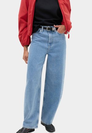Person iført lyseblå vidde jeans, sort bælte, sort top, rød jakke og sorte sko, står med den ene hånd i lommen.