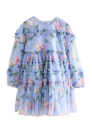 Niña con el cabello largo y oscuro, vestida con un vestido azul claro con flores y volantes y zapatillas blancas, de pie sobre un suelo claro contra un fondo liso.
