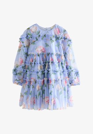 Mädchen mit langen dunklen Haaren, das ein hellblaues Blumenmusterkleid mit Rüschen und weißen Sneakers trägt, steht auf einem hellen Boden vor einem schlichten Hintergrund.
