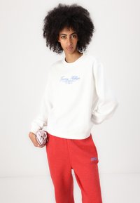 Hanorac alb cu logo-ul albastru "Tommy Hilfiger", cu manșete din tricot, asortat cu pantaloni de trening roșii cu un logo similar. Modelul ține o bentiță roz.