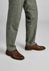 Bruine leren schoenen met een ronde neus, veters en een gestructureerd oppervlak, gecombineerd met grijsgelakte broek, zichtbaar vanaf het onderbeen.