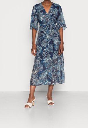 Kobieta ubrana w granatową sukienkę kopertową z białymi wzorami liści, o długości do połowy ramienia, oraz białe sandały na obcasie z plecionymi paskami.