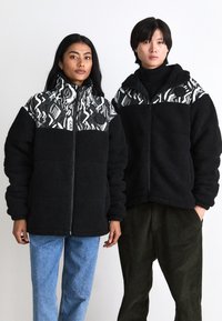 Black fleece jackets with abstract monochrome upper patterns, featuring high collars, zip closures, and a relaxed fit, worn with casual pants.