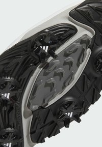 Gros plan d'une semelle cramponnée noire et grise avec plusieurs pointes arrondies et motifs texturés antidérapants sur une chaussure de sport.