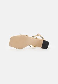 Gold metallic high-heeled sandal with a smooth beige sole and black rubber heel. Features crisscross straps, highlighting an open design.