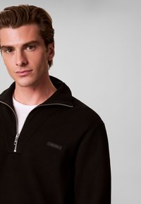 Jeune homme aux cheveux châtain clair portant un pull noir zippé Calvin Klein par-dessus une chemise blanche, sur un fond gris uni.