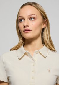 Polo beige con colletto classico, pattina con tre bottoni e un piccolo logo ricamato sul lato sinistro. Tessuto morbido e strutturato.