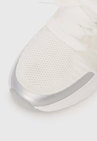 White athletic shoe with a textured mesh upper, silver toe cap, and white laces. Features a rounded sole with subtle design elements.