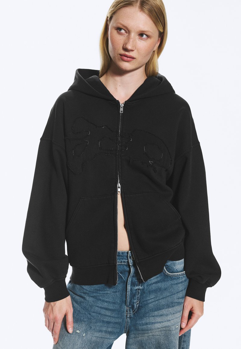 Jeune femme portant un sweat à capuche noir zippé avec un design subtil, associé à un jean bleu, regardant légèrement sur le côté sur un fond uni.