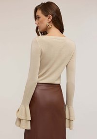 Top beige a maniche lunghe con polsini a campana, realizzato in un materiale morbido e testurizzato. Abbinato a una gonna in pelle marrone.