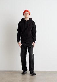 Melna kapuce ar auklītēm, mazs logo, kombinēta ar melnajām džinsām un melnajām kurpēm. Modelis nēsā spilgti sarkanu bārkstī, stāvot pret neitrālu fona.