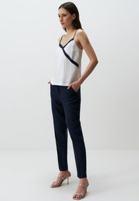 Witte mouwloze top met een diagonale marineblauwe strap, gecombineerd met marineblauwe broek en zilveren gespenhakken. Glad materiaal met een relaxte pasvorm.