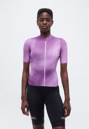 Asmuo, dėvintis prigludusią violetinį gradientinį dviračių sporto marškinėlį su trumpomis rankovėmis ir juodus dviračių šortus, stovintis prieš paprastą baltą foną.