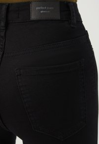 Juodos aukštu liemeniu džinsai su viena užpakaline kišene ir odiniu etiketu, ant kurios užrašyta "puikūs džinsai gina tricot" ant liemens.