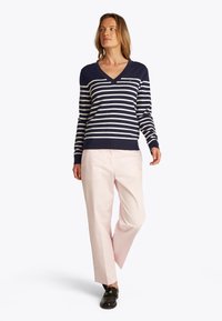 Kobieta w granatowo-białym swetrze w paski z dekoltem w serek, jasnoróżowych spodniach i czarnych mokasynach, stojąca z jedną nogą założoną na drugą.