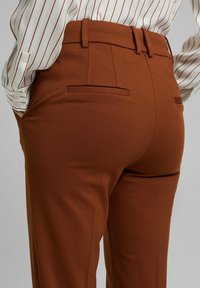Hnědé zakázkové kalhoty s hladkou texturou, plochými zadními kapsami a přiléhavým pasem. Látka má mírný lesk.