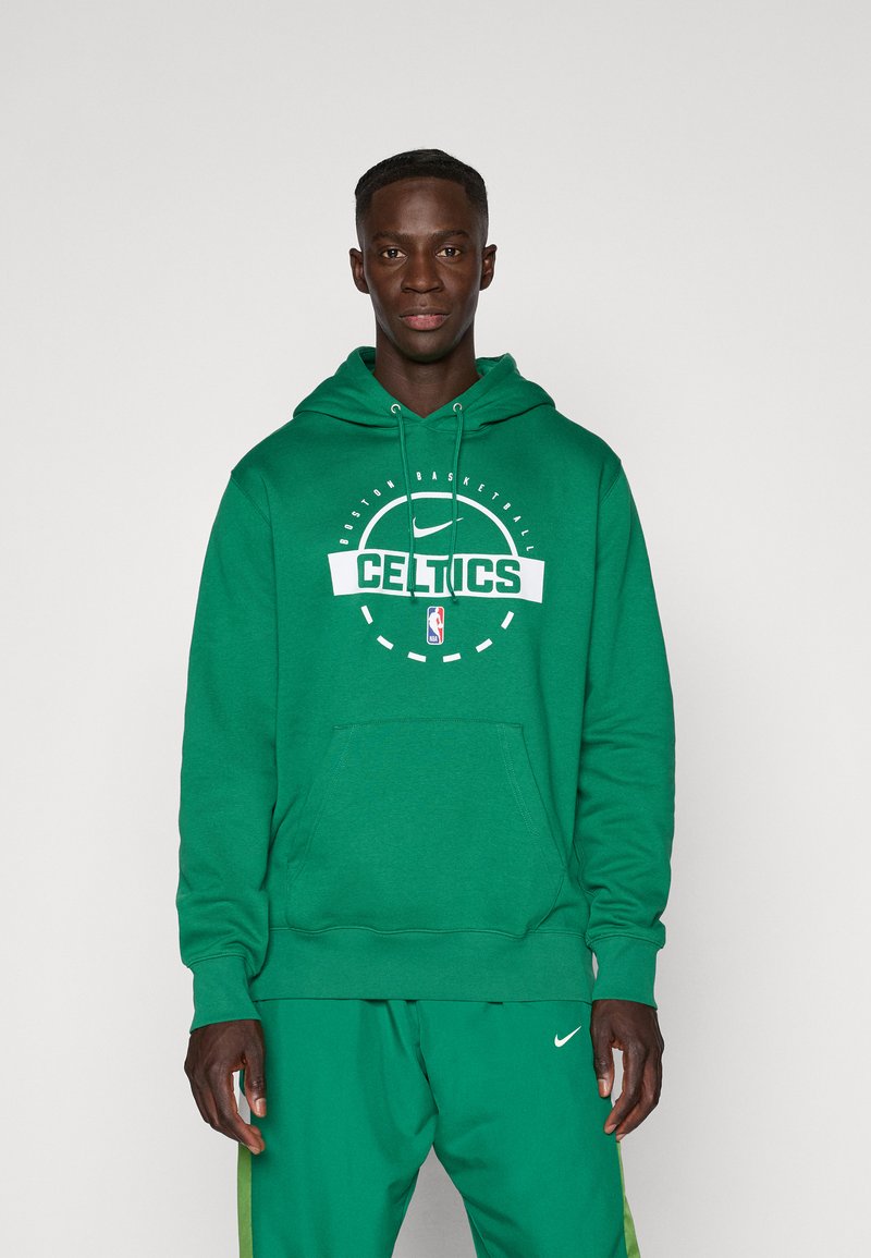 Homme portant un sweat à capuche vert des Boston Celtics et un pantalon vert assorti avec des logos Nike, debout devant un fond clair uni.