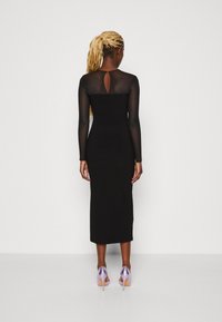 Czarna, dopasowana sukienka midi z długimi przezroczystymi rękawami, z detalem w formie dziurki z tyłu oraz fioletowymi szpilkami. Gładka, elegancka struktura materiału.