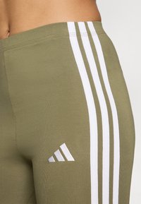 Leggings verde oliva con design a tre strisce bianche sul lato, caratterizzati da un piccolo logo Adidas argentato vicino all'orlo. Texture liscia.