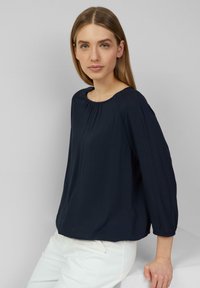 Marineblaue Bluse mit Rundhalsausschnitt, dreiviertellangen Ärmeln und dezenten Falten auf der Vorderseite; der Stoff wirkt weich und leicht.