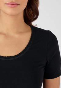 Top nero a maniche corte con scollatura a scallop e cuciture semplici, realizzato in materiale morbido e elasticizzato. Inquadratura ravvicinata del colletto.